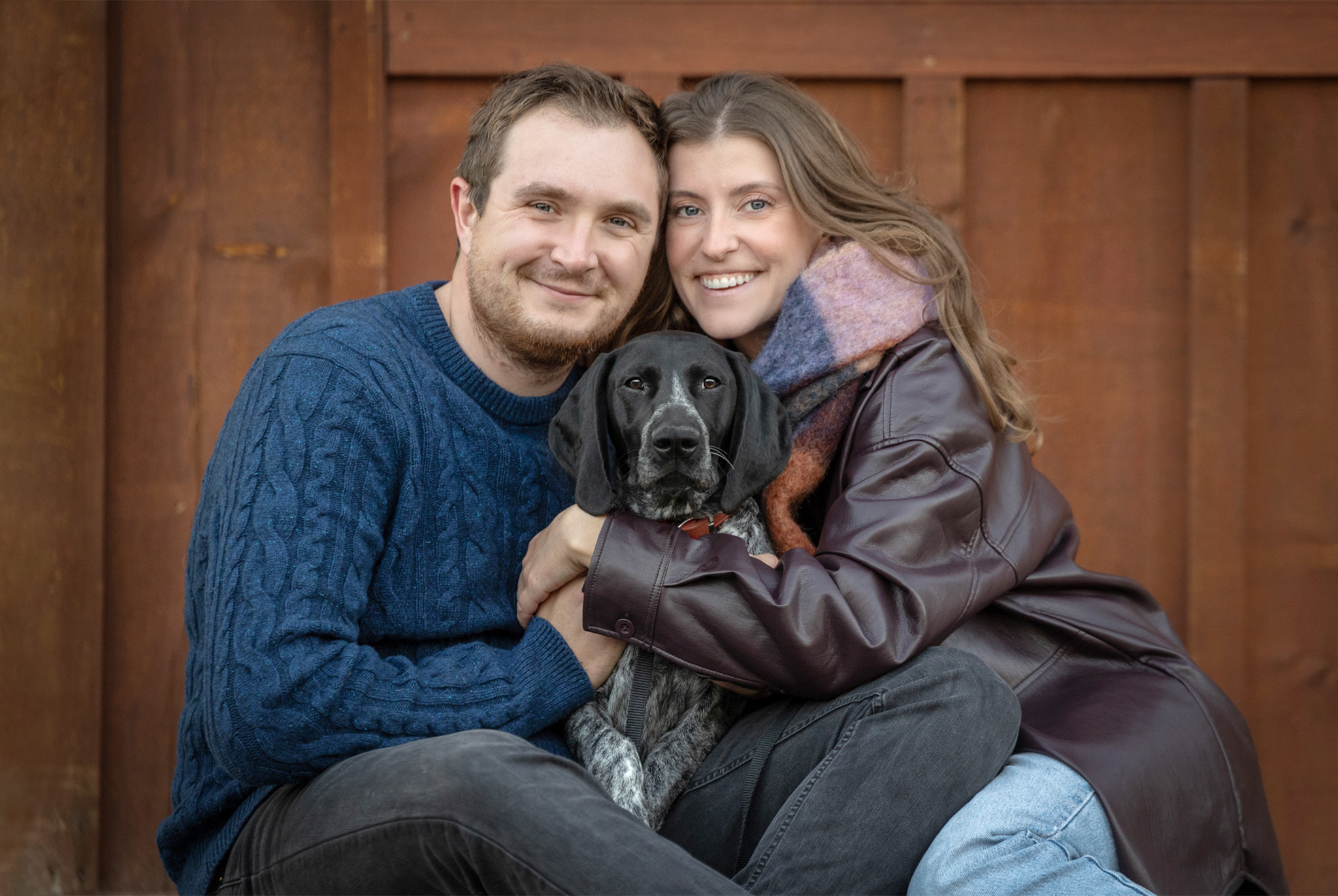 Young couple wearing fall clothes against wood wall hugging their pointer and smiling